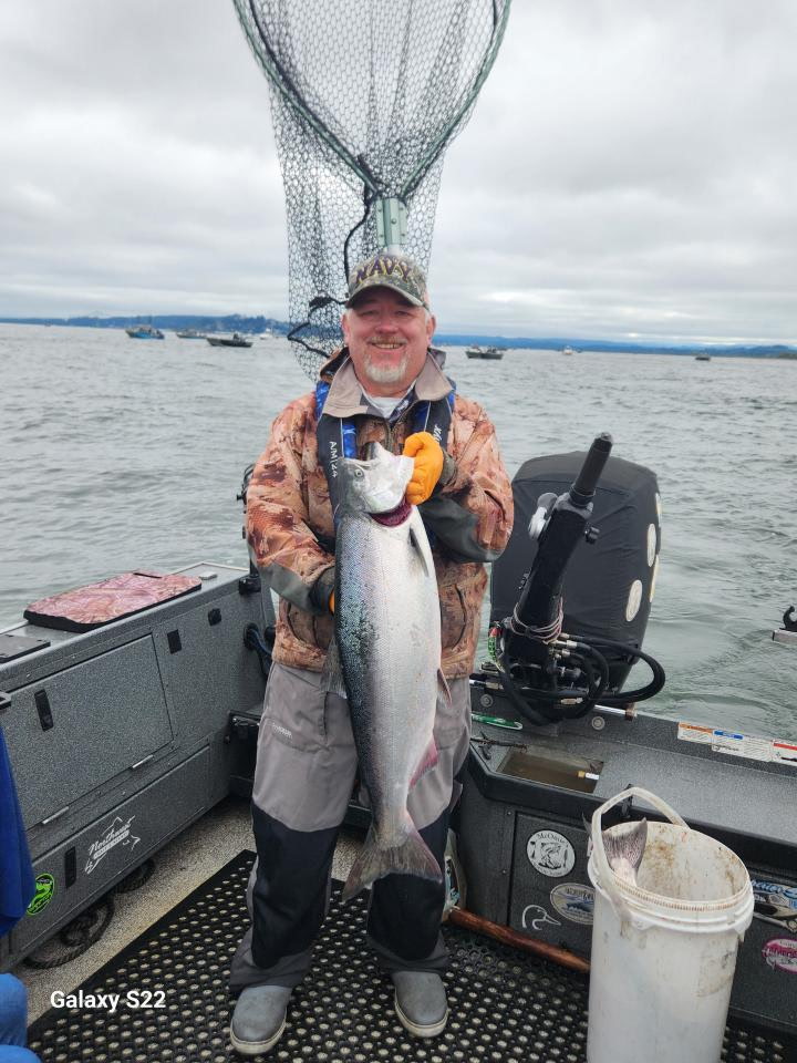 Tony Owsian on his guided fishing boat holding a fresh Chinook salmon, Navy ball cap, Columbia River estuary with the Buoy 10 fishing armada in the background.