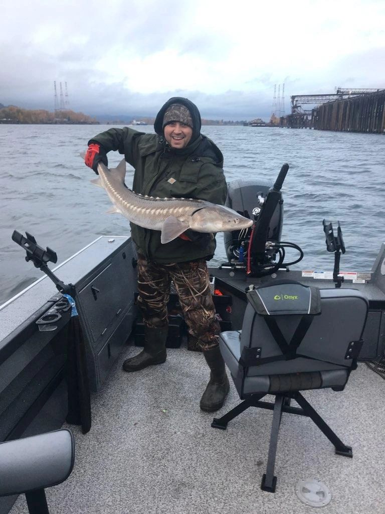 A client in heavy rain gear holds a Columbia River white sturgeon, industrial port and gantry cranes in the background.
