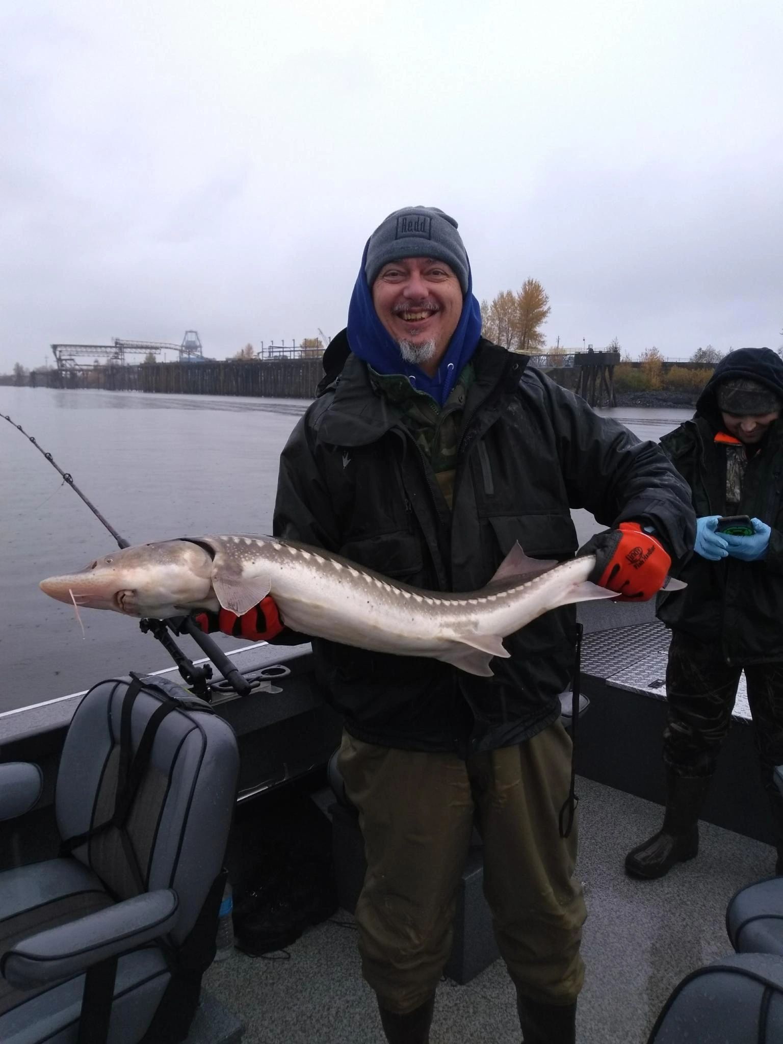 A bearded angler in a beanie and rain gear holds a Columbia River white sturgeon on a misty fall day.