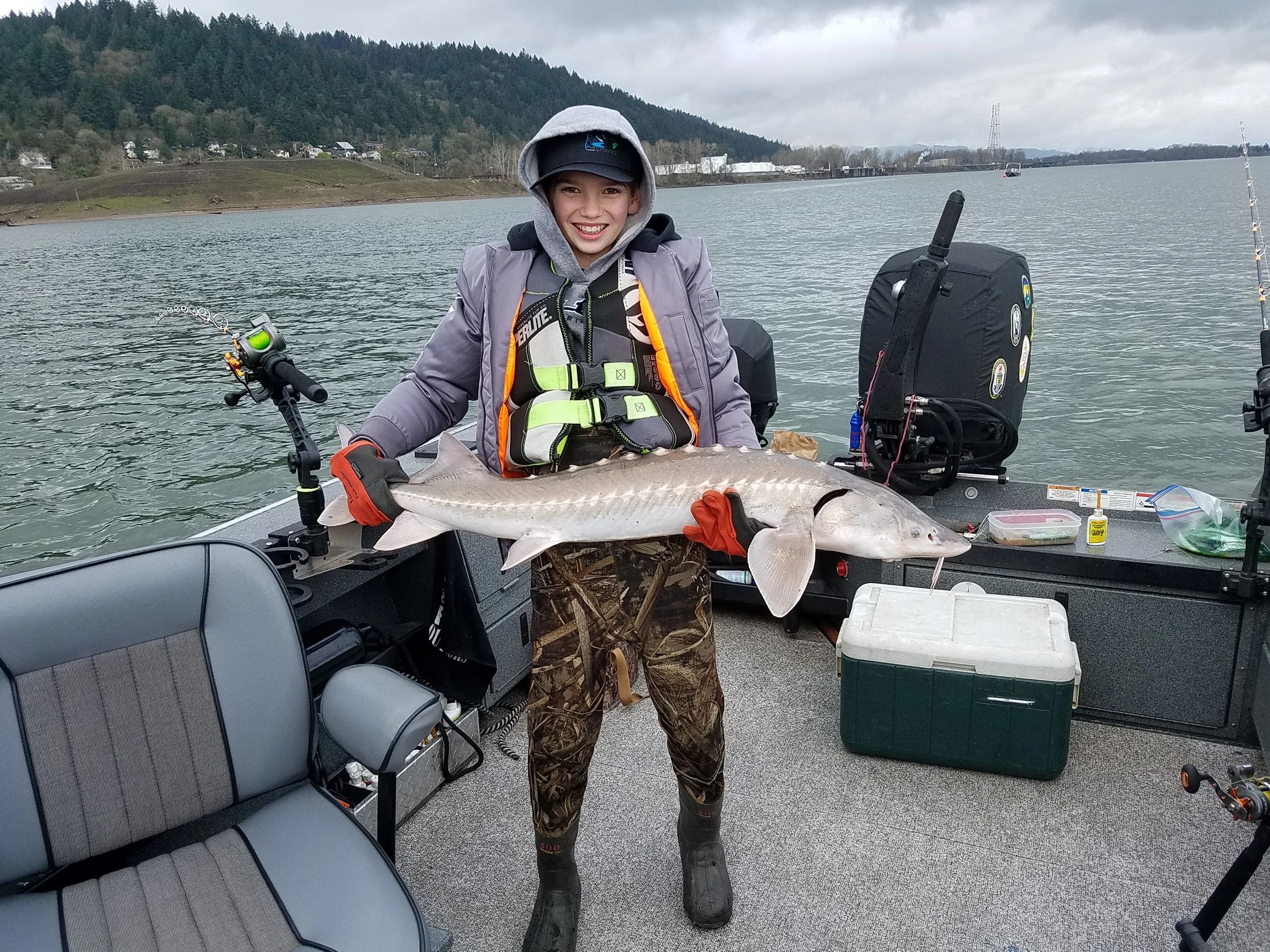 A young angler in winter gear holds a Columbia River white sturgeon on Tony's boat, forested hills in the background.