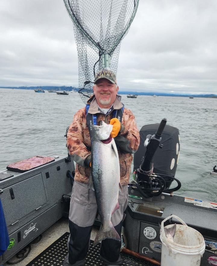 Tony Owsian on his guided fishing boat holding a fresh Chinook salmon, Navy ball cap, Columbia River with other boats in the background.